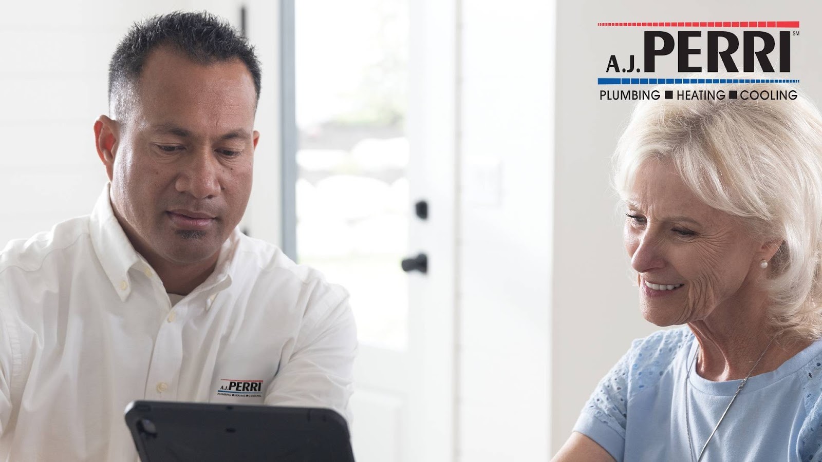 A technician from A.J. Perri Plumbing, Heating, and Cooling is showing a customer information on a tablet, both smiling and engaged in a discussion.